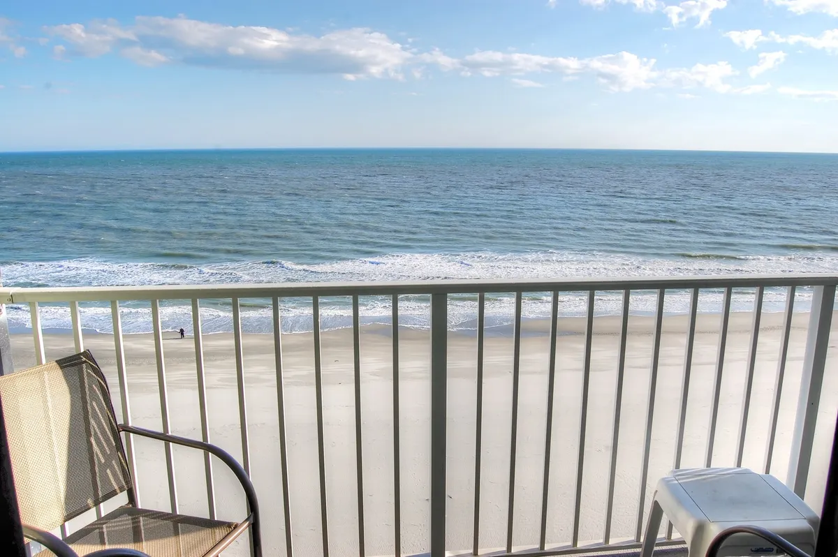 The hero image showcases a private balcony at Sands Beach Club 916, complete with comfortable chairs overlooking endless ocean views and soft sandy beaches below. The sun is rising over the Atlantic, casting a warm, serene glow across the water and inviting guests to relax, read, or simply soak in the peaceful sounds of the waves.