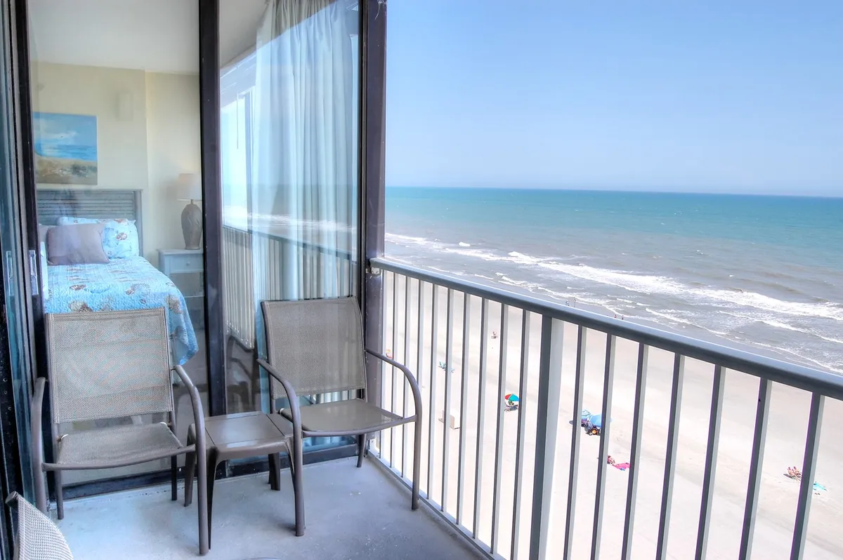 A private balcony overlooking the sparkling ocean and sandy beach, furnished with chairs for lounging. The view is serene, with gentle waves and the open sky, inviting guests to relax, sip coffee, or share sunset moments steps from the living room.