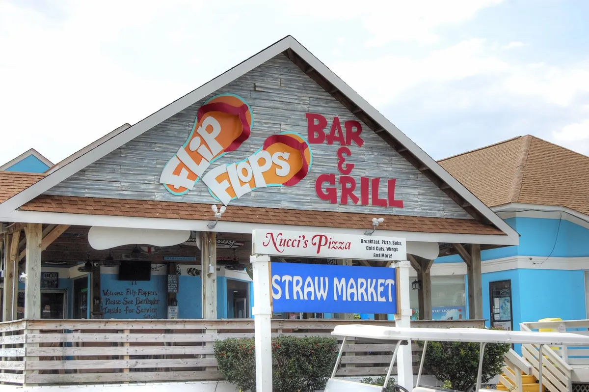 The exterior of a lively local marketplace featuring the Flip Flops Bar & Grill, Straw Market, and Nucci's Pizza signs, with an inviting and casual atmosphere ideal for dining, shopping, and socializing.