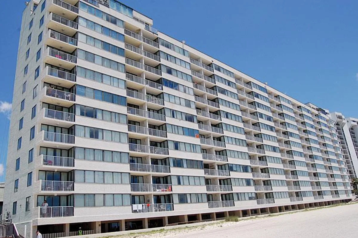 A contemporary exterior shot of the condo building, featuring private balconies, large windows, and a modern facade. Residents are seen relaxing on the balconies, enjoying panoramic beach views.