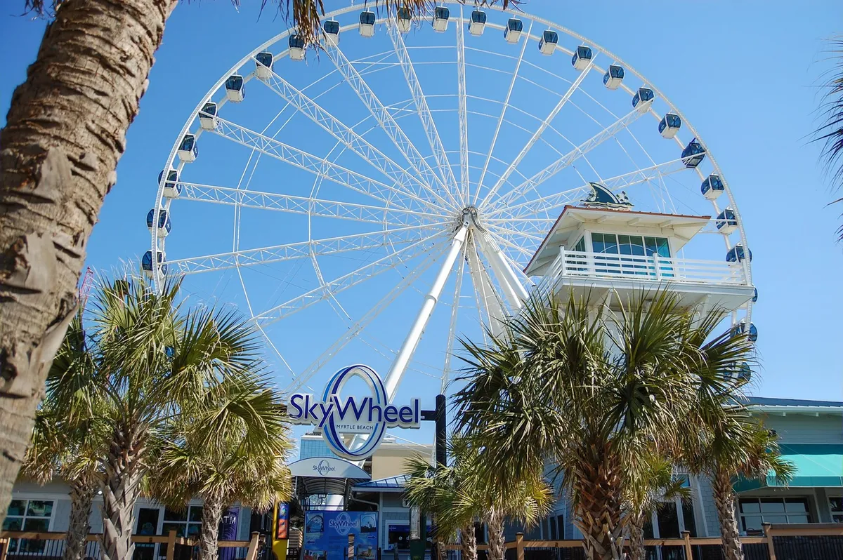 The image features the iconic Myrtle Beach SkyWheel, entrance building, and palm trees, with a cheerful mood. It suggests excitement and memorable experiences, with the SkyWheel offering breathtaking views of the coastline and being a must-visit for visitors.
