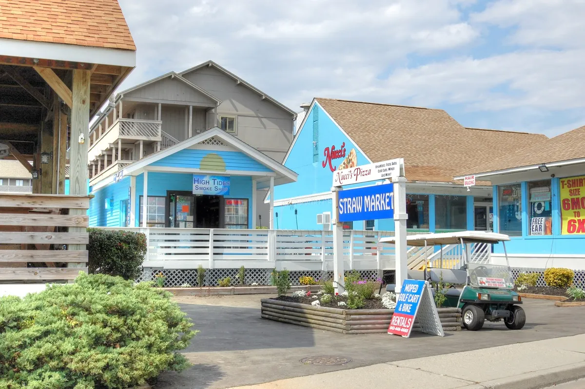 A cheerful scene of the Straw Market area, showcasing colorful blue buildings and signs for Nina's Pizza and High Tide Smoke Shop, with visitors enjoying the vibrant outdoor setting.