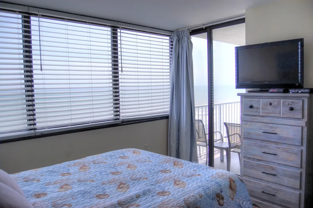 A spacious bedroom featuring a comfortable bed, large windows revealing a stunning ocean view, a dresser, and a television. The room is filled with natural light and exudes a tranquil, peaceful mood.