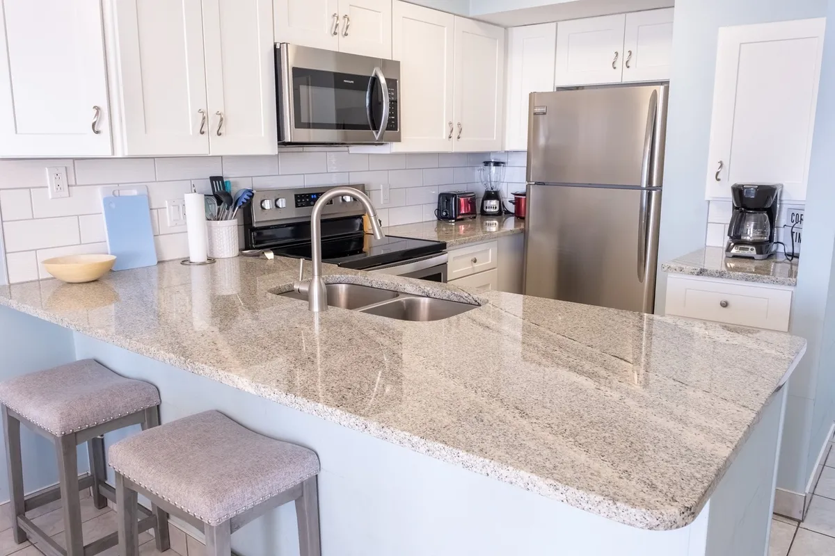 A warm, welcoming kitchen scene featuring a granite countertop, stainless steel appliances, and bar stools. The layout encourages socializing and casual family meals, with space for guests to gather around the kitchen island. The overall mood is cozy yet lively, reinforcing the idea of togetherness.