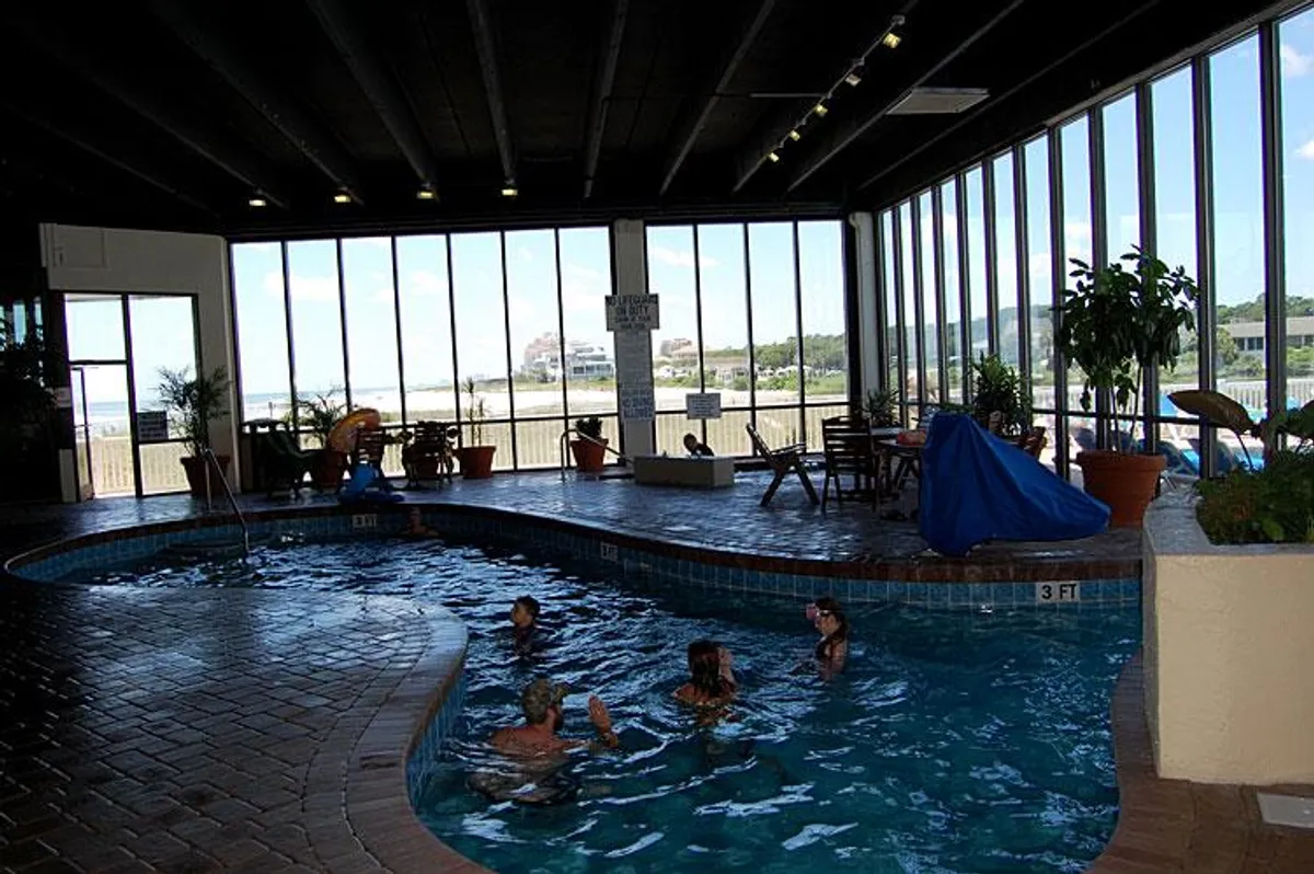 A bright indoor swimming pool with large glass windows letting in plenty of natural light. People are enjoying the pool, lounging on the deck, and indoor plants add a touch of lushness. The mood is joyful, inviting guests to swim, relax, and socialize.