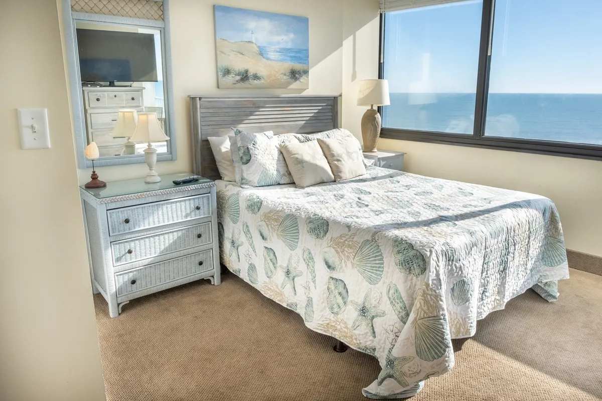 A bedroom with a shell-patterned quilt on the bed, large windows offering an ocean view, a side table with a lamp, and a mirror reflecting natural light. The atmosphere is calm and inviting, emphasizing relaxation and comfort.