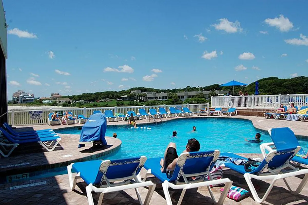 An outdoor swimming pool surrounded by lounge chairs and umbrella-covered seating. Guests are swimming, sunbathing, and relaxing in a leisurely, inviting atmosphere. The setting is ideal for both play and relaxation.