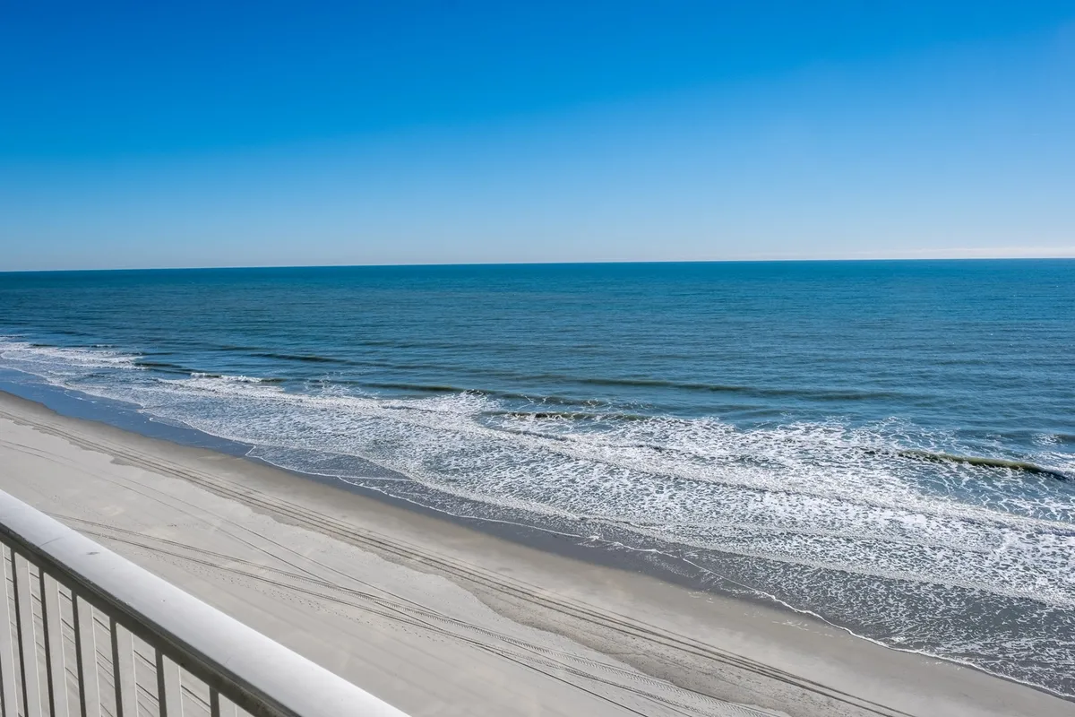 A wide, unobstructed view of the ocean waves gently washing onto a sandy beach under a clear blue sky. The composition emphasizes the expansive shoreline and tranquil, natural beauty of Myrtle Beach at sunrise or sunset.
