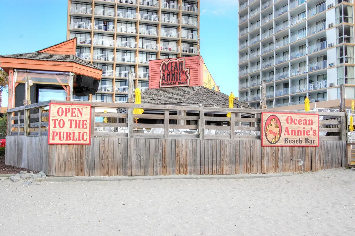 Ocean Annie's Beach Bar exterior with an inviting open-to-the-public banner, wooden enclosure, and yellow umbrellas set against a coastal backdrop, creating a relaxed, social gathering spot by the beach.