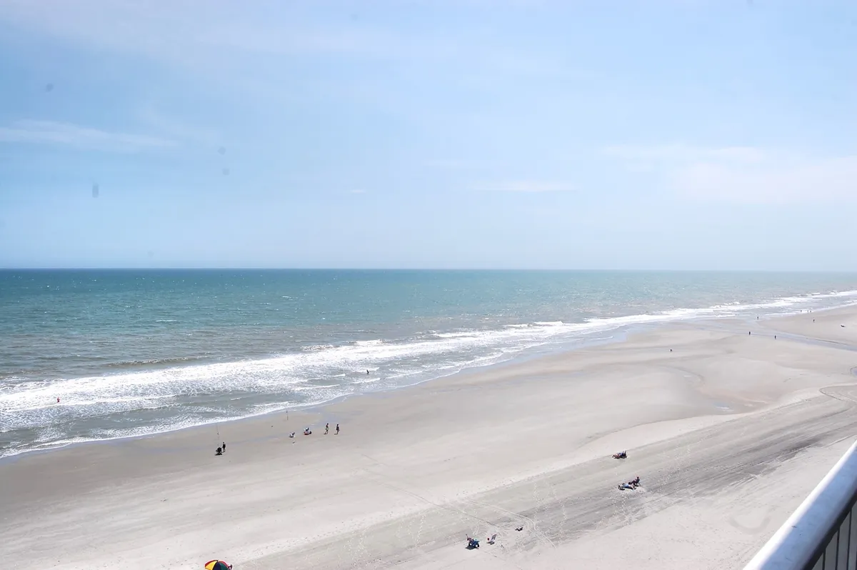 This image shows a sweeping view of the Atlantic Ocean, sandy beach, and distant shoreline with people leisurely walking along the shore. The inviting coastline captures the essence of direct beach access and the natural beauty families can enjoy together.