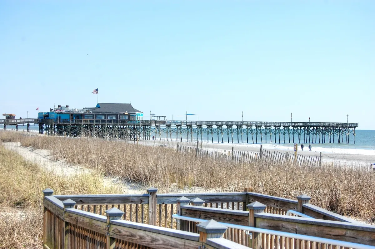 A peaceful exterior shot of a pier stretching over the ocean, framed by sand dunes and a wooden boardwalk leading to the water. The tranquil scene invites leisurely walks, fishing, and group relaxation along the shore.