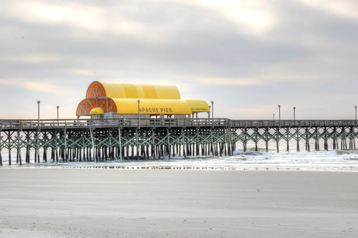 A wide view of Apache Pier with its distinctive yellow roof structure and strong pier supports stretching into the ocean. The pier area is lively and ideal for walking, sightseeing, and starting a beachside adventure.